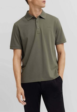 Uomo che indossa una polo verde oliva a maniche corte con colletto e bottoni, abbinata a pantaloni neri, in piedi davanti a uno sfondo neutro.