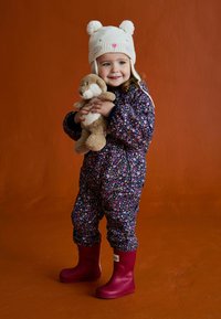 Kolorowy kombinezon w kwiatowy wzór na ciemnoniebieskim tle, zestawiony z różowymi gumowymi butami i kremową czapką z motywem misia. Miękki miś w ramionach.