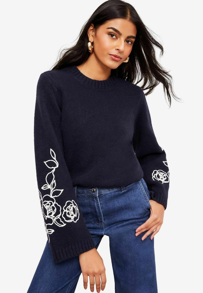 Pulover bleumarin cu guler cu nervuri și modele brodate cu trandafir alb pe mânecile clopot, asortat cu blugi. Textură moale.