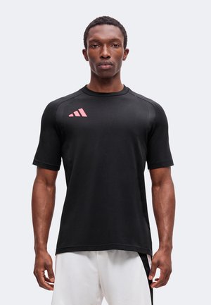 Homme portant un t-shirt de sport noir à manches courtes avec un petit logo rose sur la poitrine et un short blanc avec une bande noire.