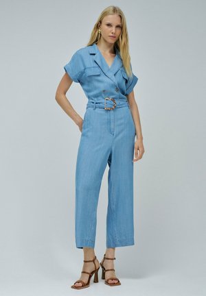 Une combinaison en denim bleu clair avec des manches courtes, une taille ceinturée avec une boucle ronde et un pantalon à jambes larges. Le tissu a une texture lisse.