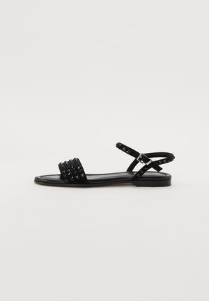 Sandale noire avec détail de sangle cloutée, semelle plate et sangle de cheville réglable. Fabriquée en matériau texturé avec un design épuré et minimal.