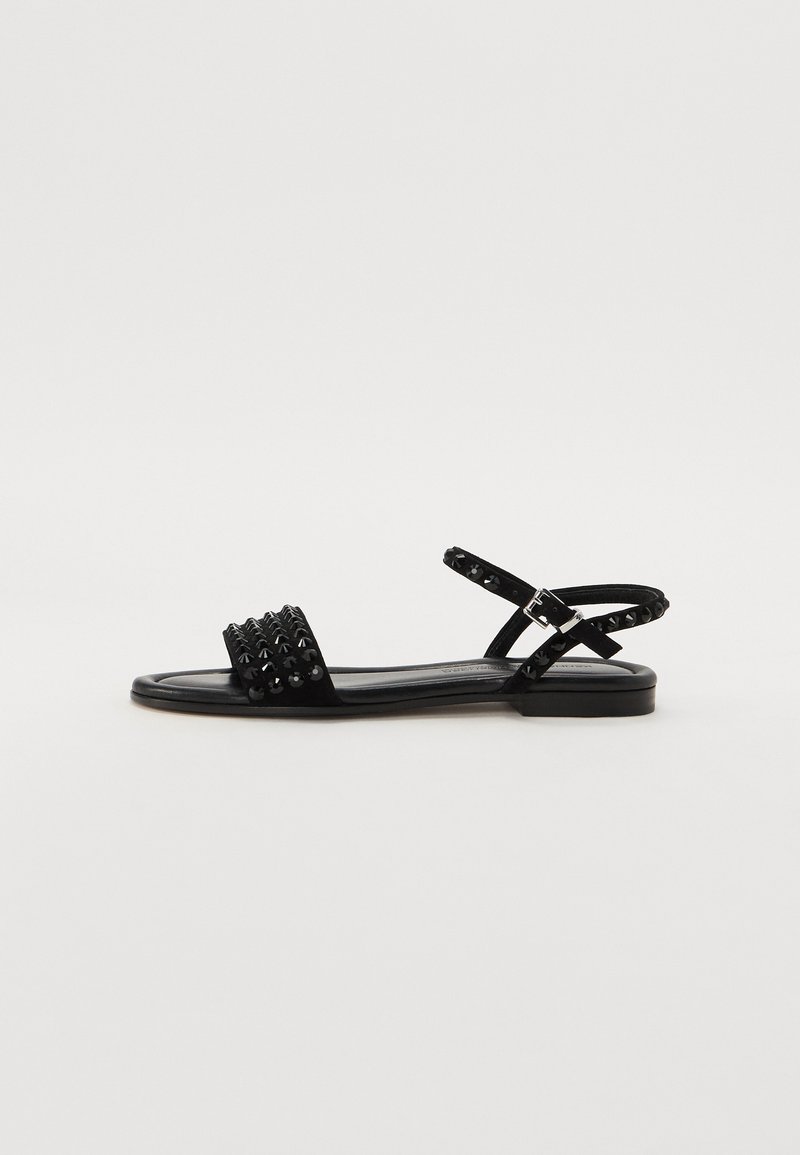 Svart sandal med naglede detaljer på stroppen, flat såle og justerbar ankelrem. Laget av teksturert materiale med et elegant, minimalistisk design.
