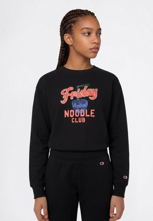 Unge kvinde med fletninger iført en sort sweatshirt med teksten "Friday Noodle Club" og en blå skålgrafik, kigger til siden.