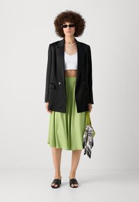Svart oversized blazer, hvit crop topp, flytende limegrønn skjørt, svarte høye sandaler, holder et mønstret skjerf. Tilbehøret inkluderer hoopøredobber.