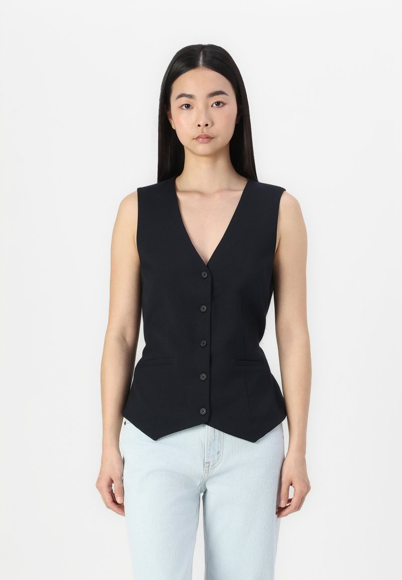 Black sleeveless vest with a V-neck, featuring six buttons and two front pockets. Shaped hem and smooth fabric. Worn over light blue jeans.