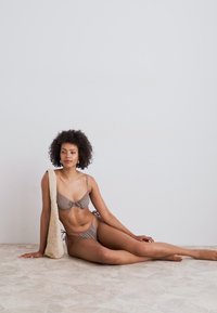 Brun bikiniuppsättning med en knuten topp och knytbara bottnar, i kombination med en texturerad beige klädsel, på ett neutralt kaklat golv.