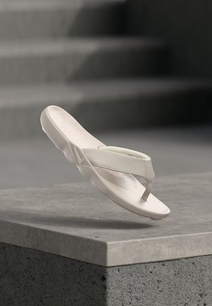 Valge Nike varbavahe-sandaal hõljub halli betoonserva kohal, taustal ähmased trepid.