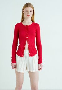 Κόκκινο ριμποτό μπλουζάκι μακρυμάνικο με χρυσά κουμπιά, συνδυασμένο με χαλαρά, κρεμ και ριγέ σορτς. Λείο ύφασμα, εφαρμοστή σχεδίαση.