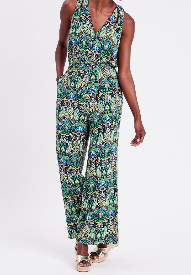 Combinaison à motifs vert, bleu et noir avec un décolleté en V, des jambes larges et une taille cintrée. Associée à des sandales à plateforme dorées.