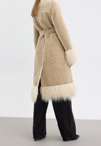 Lungo cappotto beige con un tessuto effetto texture, trim in pelliccia artificiale all'orlo e alle maniche, e una cintura da annodare. Esposto di spalle su uno sfondo neutro.