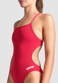 Maillot de bain rouge avec de fines bretelles et un design découpé, arborant un logo blanc sur le côté. Fabriqué en tissu lisse et extensible.