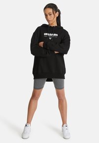 Sudadera negra de gran tamaño con texto gráfico blanco, combinada con pantalones cortos de ciclismo grises y zapatillas blancas. Tejido suave, corte relajado y cordón.