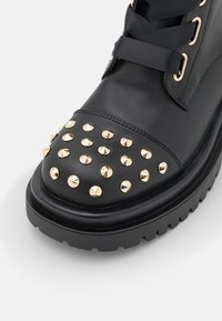 Botte en cuir noir avec des accents cloutés dorés sur le bout et une semelle épaisse noire. Comprend des lacets décoratifs noirs et des éléments en métal.