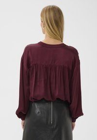 Burgundy blouse with a round neck, gathered back detail, and long sleeves. Paired with a black leather skirt featuring a central zipper.