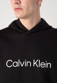 Juoda palaidinė su kengūros kišenėmis, reguliuojamu raišteliu ant kapšo ir priekyje esančiu baltu logotipu "Calvin Klein". Lygus audinio paviršius.