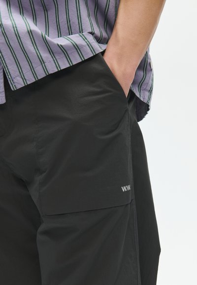 Άτομο που φοράει μαύρα cargo σορτς με το χέρι στην τσέπη και ένα λεβαντίνη πουκάμισο με πράσινες κάθετες ρίγες.