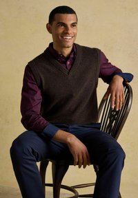 Smaidošs vīrietis, kurš valkā tumši brūnu vesti pār bordo kreklu un tumši ziliem biksēm, sēž uz koka krēsla uz bēšas fona.