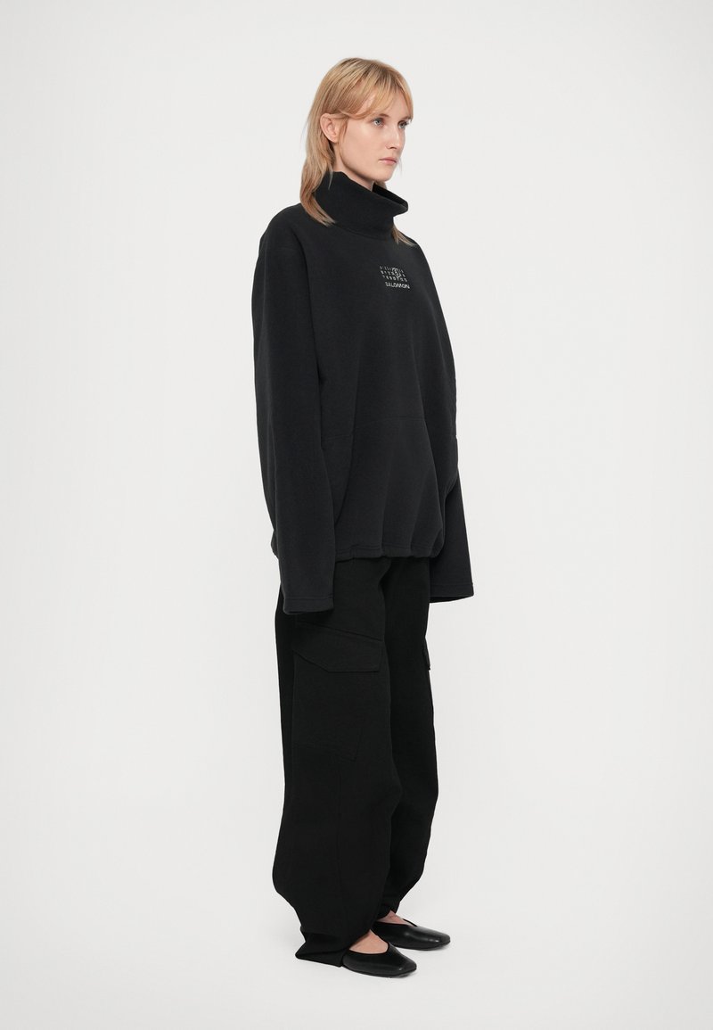 Mujer de pie, vistiendo un suéter de cuello alto negro oversized y pantalones cargo anchos negros con zapatos planos negros, frente a un fondo blanco liso.