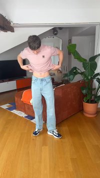 Joven con vaqueros azul claro y camisa rosa levanta su camisa, de pie en una sala de estar con un sofá marrón y una planta grande y verde cerca.