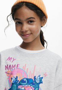 Ljusgrå sweatshirt med ribbad krage, prydd med en färgglad grafik av en tecknad figur och texten "MITT NAMN" i rosa. Mjuk tygkvalitet.