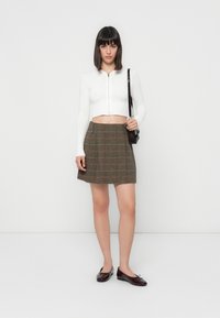 Woman standing wearing a white cropped zip-up sweater, brown plaid skirt, burgundy flats, and a matching shoulder bag, against a plain white background.