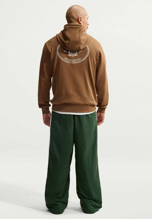 Uomo con felpa marrone con logo e pantaloni verdi a gamba larga, in piedi di spalle su sfondo semplice.