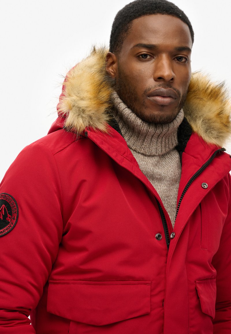 La veste parka rouge présente une capuche en fausse fourrure amovible, deux poches poitrine et un col roulé beige texturé en dessous.