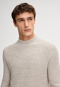 Jasno beżowy dzianinowy sweter z wysokim dekoltem typu crew, z teksturowanym wzorem i ściągaczami na mankietach. Luźny krój, miękki materiał.