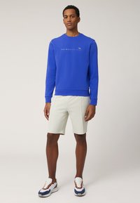 Blauwe sweatshirt met ronde hals en logoprint, gecombineerd met lichte shorts en sportieve sneakers in een mix van wit, blauw en bruine accenten.