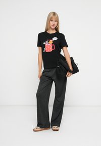 Ung kvinde iført sort fest T-shirt med en tegneserie-bjørn i en rød krus og grå vidde-bukser, mens hun holder en sort taske og står foran en hvid baggrund.