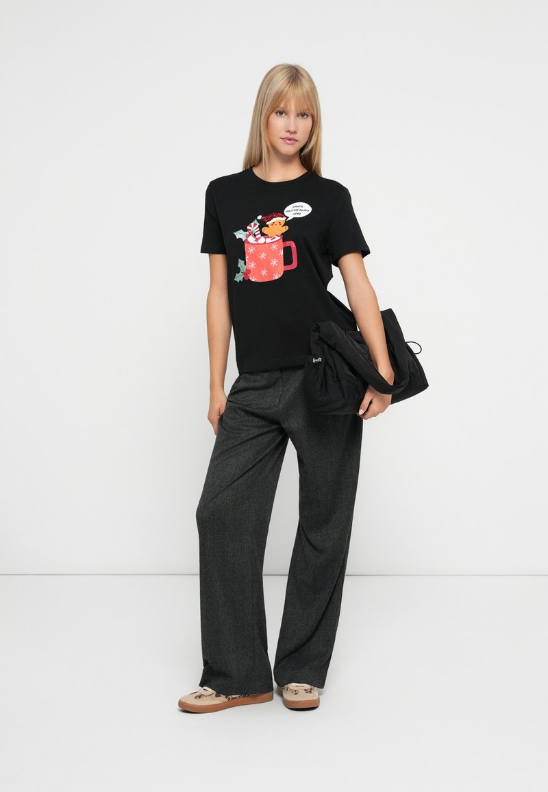 Junge Frau trägt ein schwarzes festliches T-Shirt mit einem Cartoon-Bären in einer roten Tasse und grauen weiten Hosen, hält eine schwarze Tasche und steht vor einem weißen Hintergrund.