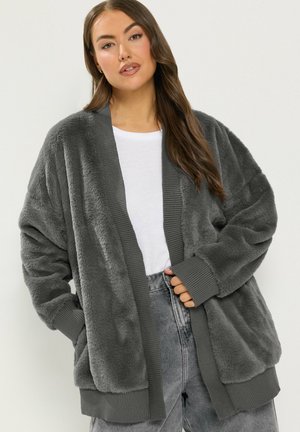 Femme portant un cardigan ouvert en peluche douce grise avec des poignets et un ourlet côtelés sur une chemise blanche et un jean gris, posant devant un fond uni.