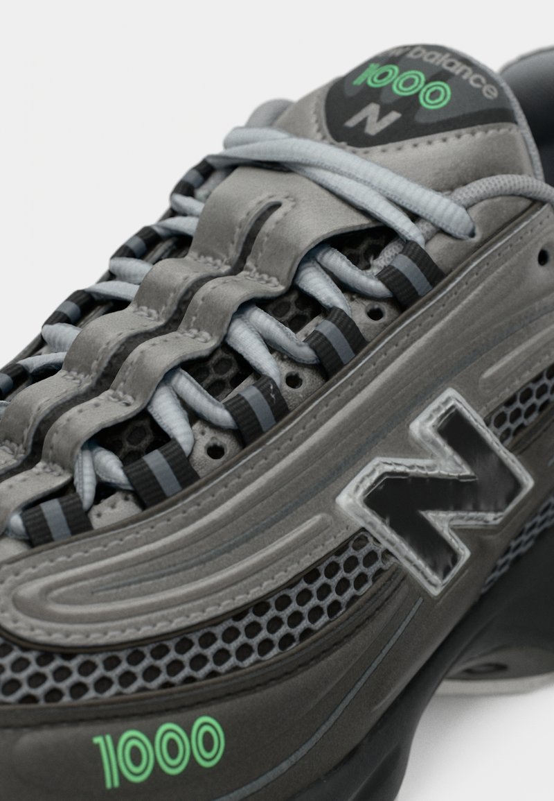 Baskets grises avec des panneaux en maille et des revêtements synthétiques texturés. Équipées d'un logo "N" noir, d'accents verts et d'un système de laçage complexe.