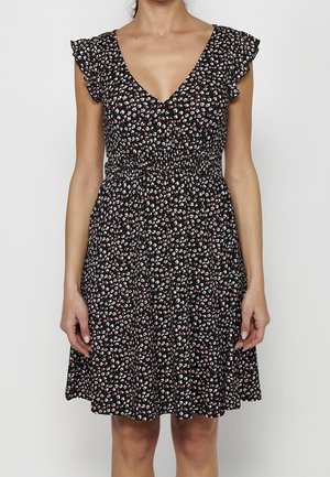 Kobieta w czarnej sukience z małym białym i różowym kwiatowym wzorem, dekoltem w serek, marszczonymi krótkimi rękawami i marszczonym detalem w talii.