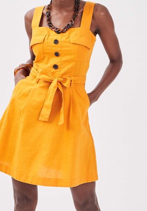 Femme portant une robe sans manches orange vif avec des boutons sur le devant, des poches poitrine, une ceinture nouée à la taille et un collier épais en écaille foncée.