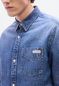 Πουκάμισο από τζιν σε ανοιχτό μπλε χρώμα με μυτερό γιακά, κούμπωμα με κουμπιά μπροστά, τσέπη στο στήθος με ετικέτα Calvin Klein σε κόκκινο και λευκό, και αντίθετη ραφή.