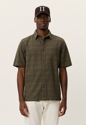 Kortærmet, olivengrøn skjorte med et struktureret, vævet mønster og knapper foran. Båret med lyse bukser og en mørk kasket.