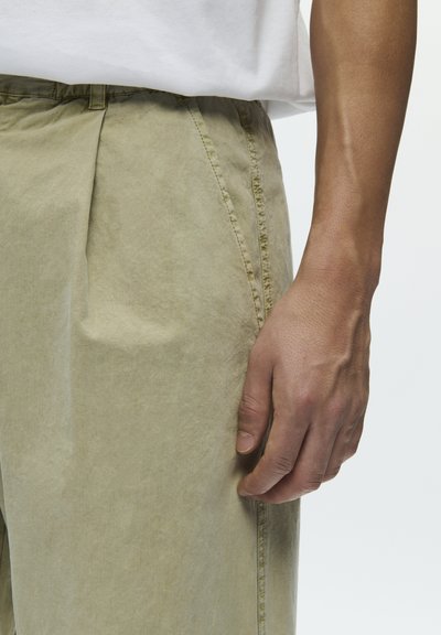 Közelkép egy személy jobb kezéről, amely bézs nadrág és fehér ing mellett nyugszik, megmutatva a bőr textúráját és a ruházat részleteit.