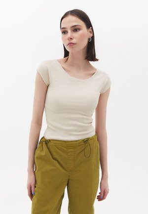 Beige eng anliegendes Rippoberteil mit halblangen Ärmeln und weitem Ausschnitt, kombiniert mit locker sitzenden olivgrünen Hose mit Kordelzug. Strukturiertes Stoffdetail sichtbar.