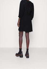 Černé šaty s nadýchanými rukávy a vyšívaným zadním dílem. Hladká látka, rozšířený tvar, nošeny s černými ponožkami a robustními loafery na podpatku.