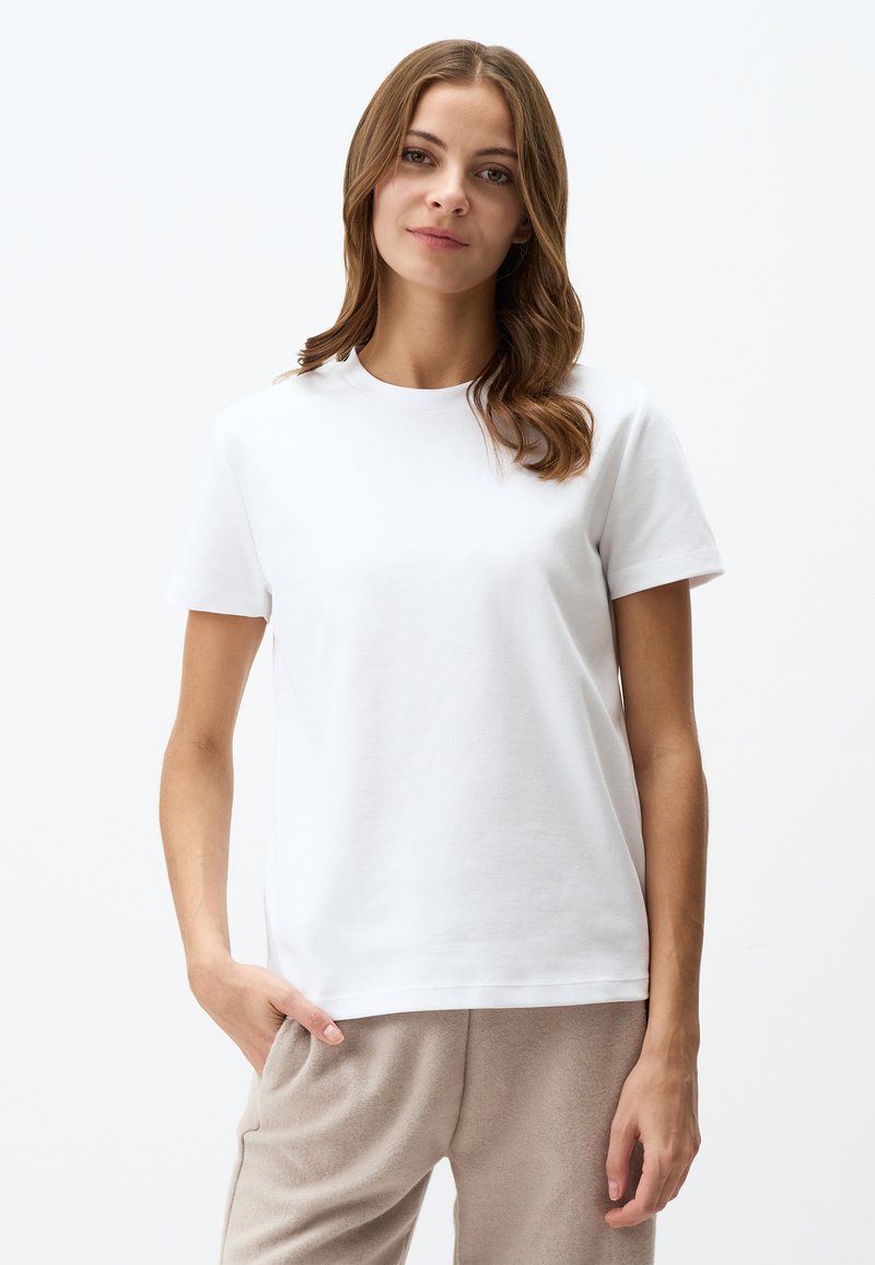 T-shirt bianco in cotone con maniche corte, scollatura rotonda, vestibilità comoda e texture morbida. Abbinato a pantaloni beige, mani infilate nelle tasche.