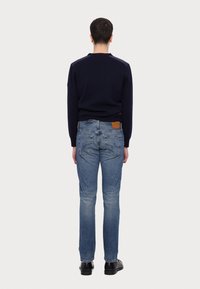 Mies, joka seisoo selin päällä tummansininen neule, vaaleansinisiä farkkuja takataskuilla ja mustia kenkiä, yksinkertaista valkoista taustaa vasten.