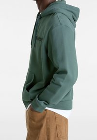 Sudadera con capucha de algodón verde con bolsillo delantero, capucha ajustable con cordón y un logotipo discreto de Vans en un tono oscuro. La modelo lleva pantalones color caqui.