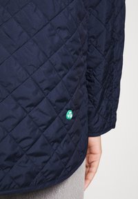 Primer plano de la manga y el lado de una chaqueta acolchada azul marino con una pequeña insignia verde y una mano visible descansando cerca de unos pantalones gris claro.