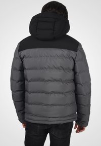 Siva puffer jakna s crnim došivkom na ramenima, s horizontalno kvadratnim dijelovima, kapuljačom i glatkom površinom. Crni naglasci na vratu i manžetama.