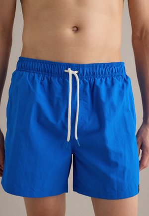 Uomo che indossa pantaloncini da bagno blu vivaci con coulisse bianca, torso e parte superiore delle gambe visibili, sfondo neutro.