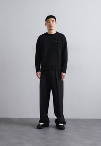 Black crewneck sweatshirt with a small logo, paired with loose-fitting black trousers and black slip-on shoes with white accents.