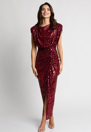 Kobieta stojąca uśmiechnięta, ubrana w bezrękawową długą suknię w ciemnoczerwonym kolorze ozdobioną cekinami, z rozcięciem z przodu i beżowymi sandałami na wysokim obcasie, na tle jednolitego szarego tła.