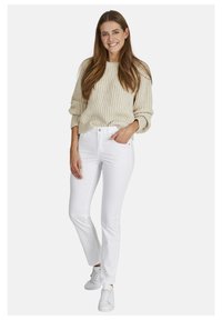 Witte katoenen jeans gecombineerd met een beige geribbelde trui. Het model draagt witte sneakers en staat met één hand op haar heup.